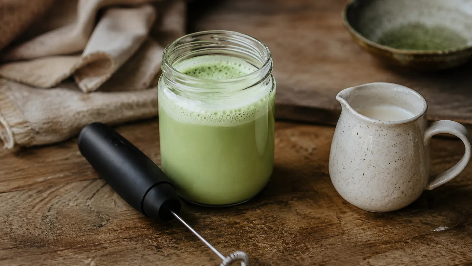 A frothy matcha latte in a glass jar next to a handheld frother on a wooden cutting board