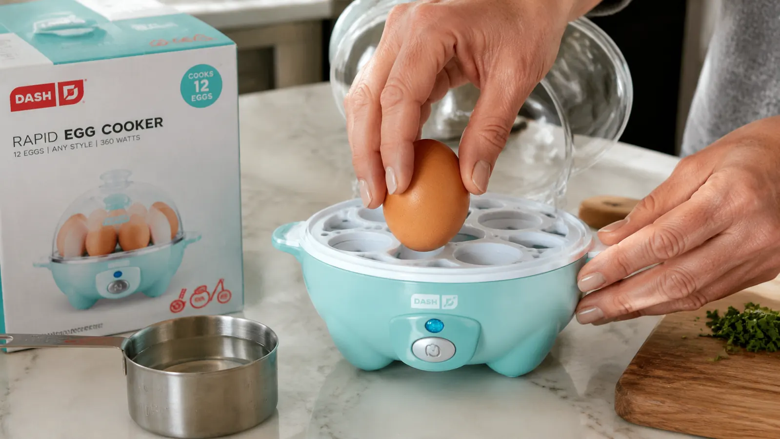 Hand placing an egg into the Dash egg cooker tray with the measuring cup of water nearby