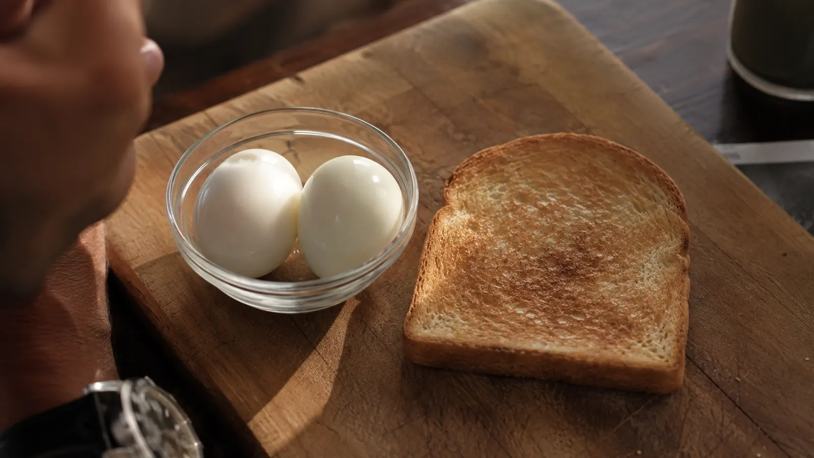 Peeled hard-boiled eggs in a small glass bowl next to toast on a wooden cutting board