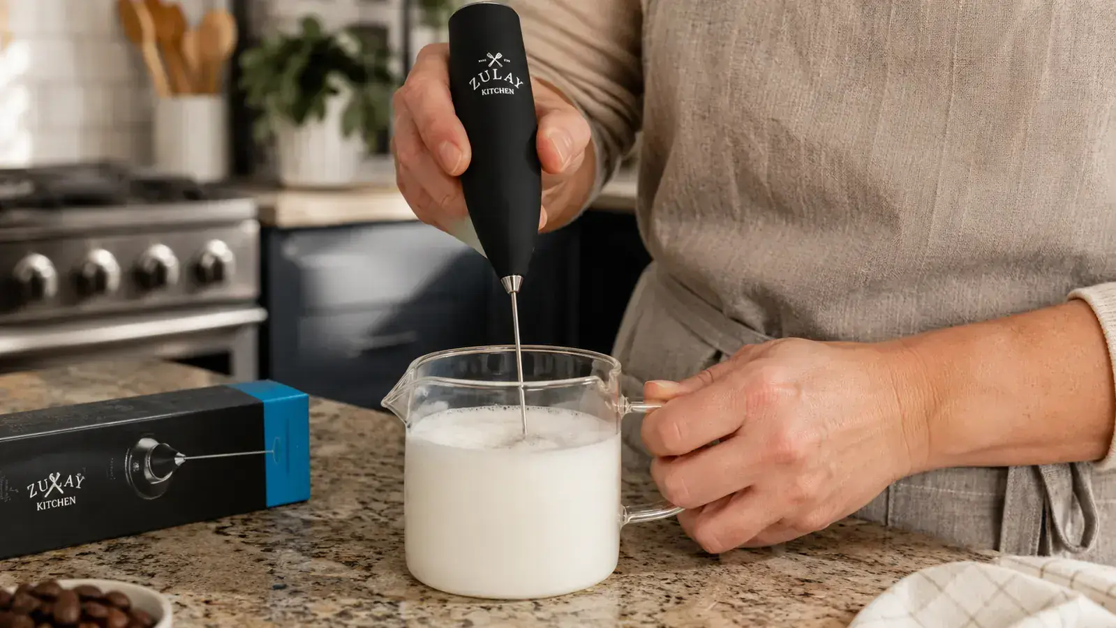 Hand holding a Zulay handheld milk frother wand submerged in a small stainless steel pitcher of milk, foam forming at the surface