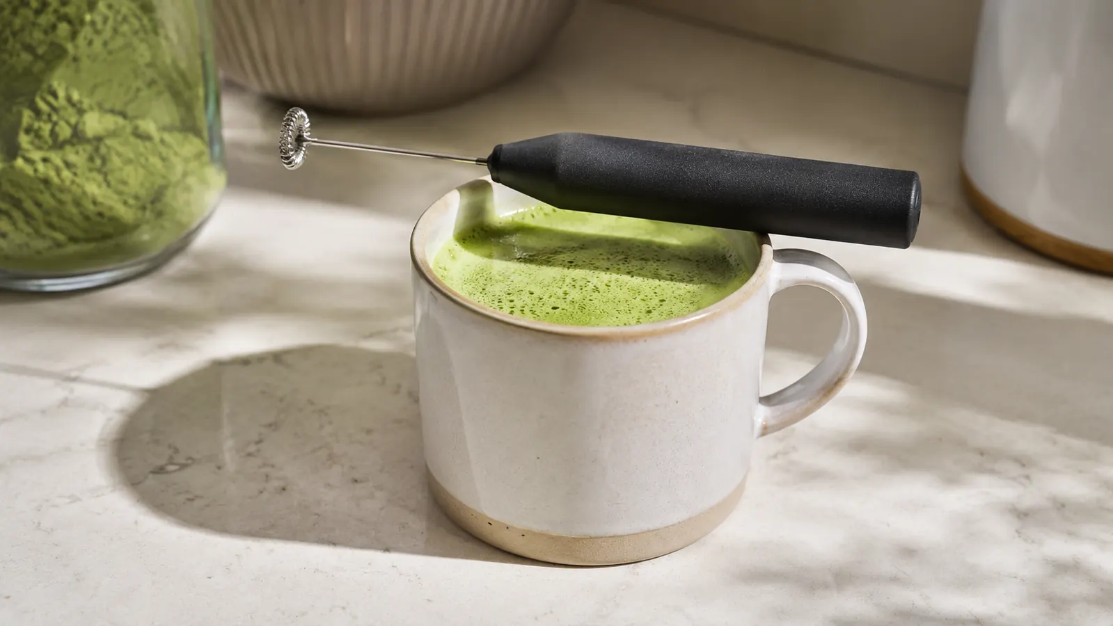 Handheld frother wand resting beside a mug of matcha latte on a sunlit kitchen counter